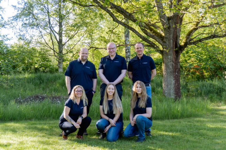 En gruppbild med träd i bakgrunden och gräs runt omkring. Tre kvinnor sitter på huk i gräset och bakom står tre män. Alla ler mot kameran och har jeans med företagstshirt på sig.
