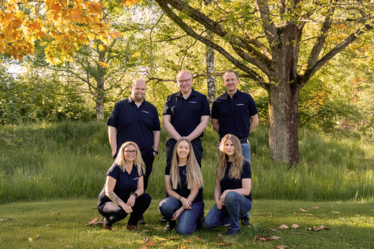 En gruppbild med träd i bakgrunden och gräs runt omkring. Tre kvinnor sitter på huk i gräset och bakom står tre män. Alla ler mot kameran och har jeans med företagstshirt på sig.