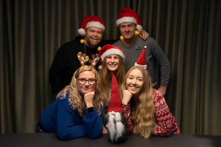 Människor som klätt upp sig juligt med tomteluvor och jultröjor. De står framför en grön gardin. En tomte i tyg syns i bild också.