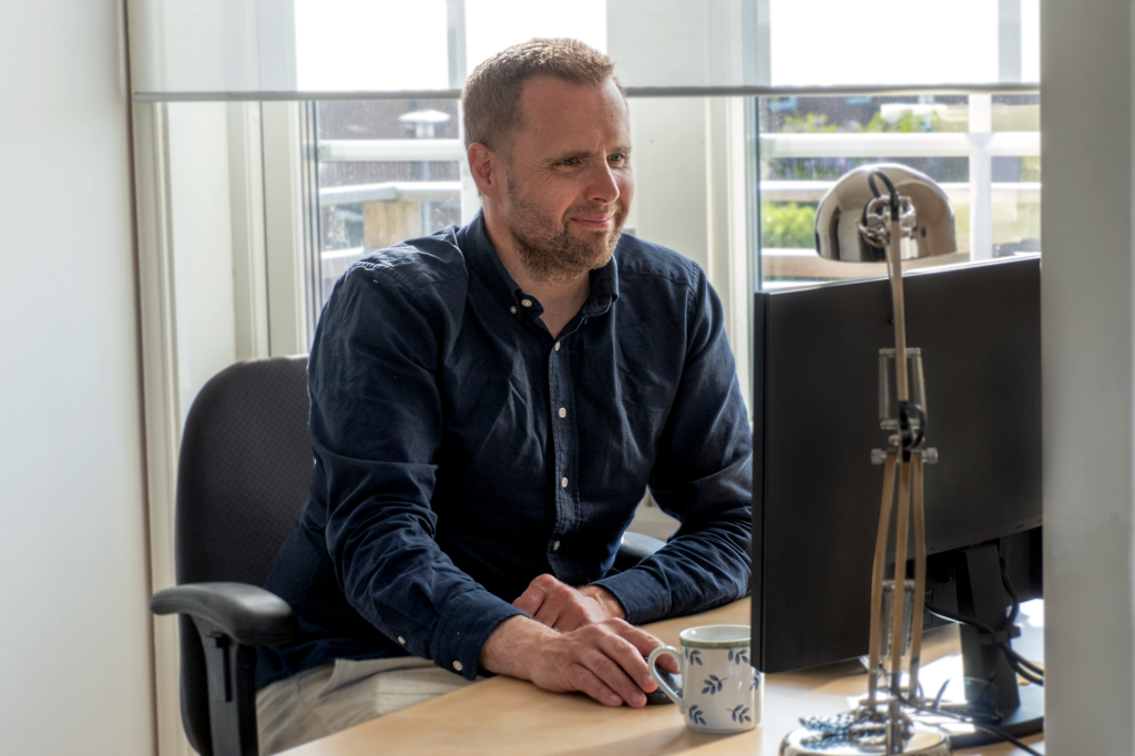 Person sitter vid ett skrivbord framför en datorskärm med en kaffekopp bredvid.