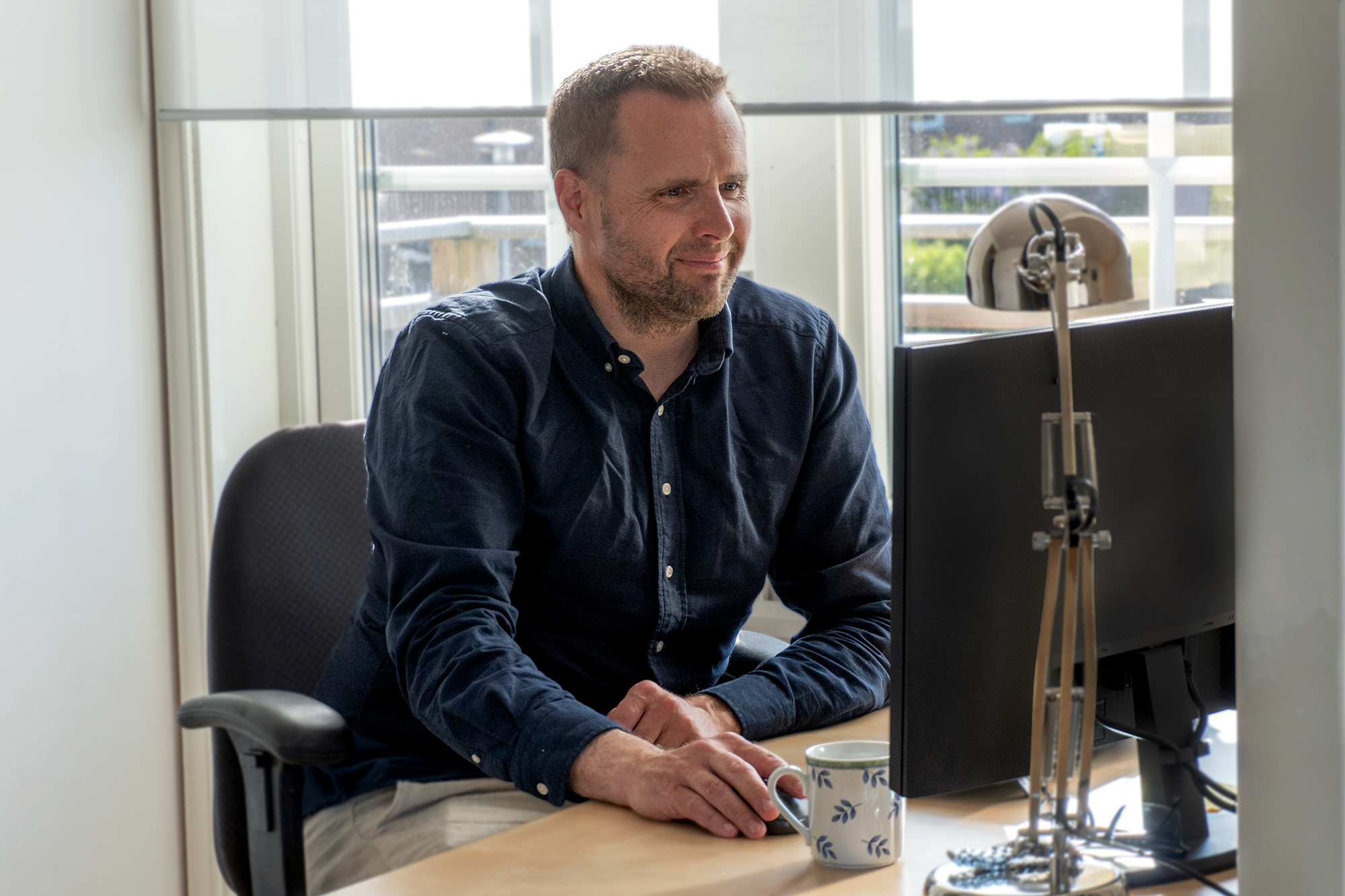 Person sitter vid ett skrivbord framför en datorskärm med en kaffekopp bredvid.