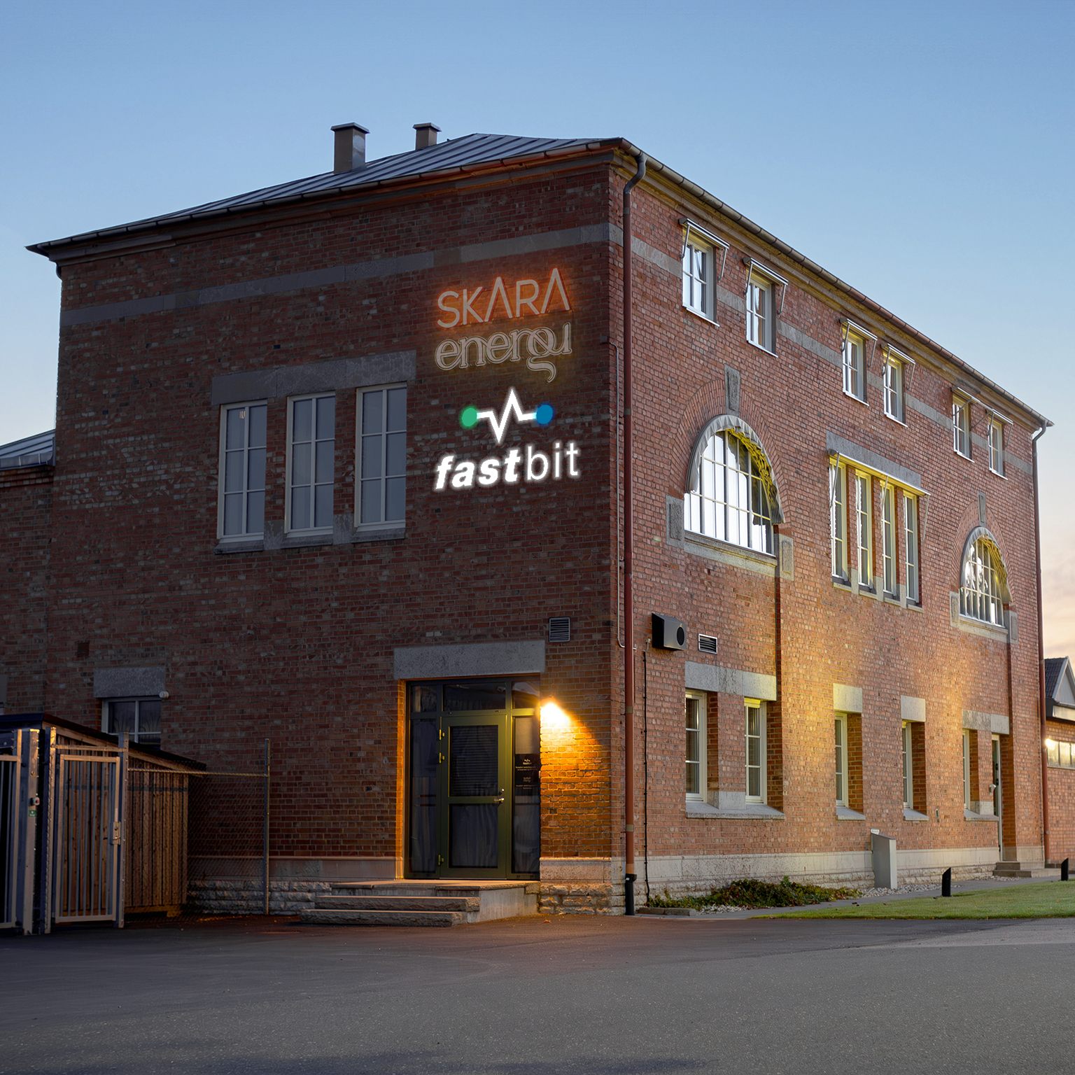 Tegelbyggnad med upplyst entré i kvällsljus. På fasaden syns skyltar med Skara Energi och Fastbit.
